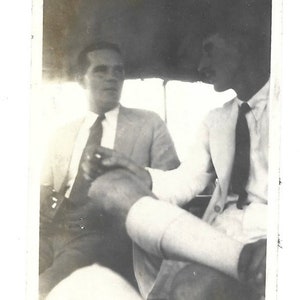 May include: Black and white photo of two men sitting in a car. The man on the left is wearing a suit and tie. The man on the right is wearing a white shirt and a dark tie.
