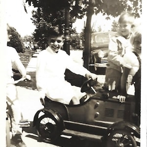 May include: A black and white photo of a young boy driving a toy car. He is smiling and wearing a white shirt. Two other children are standing behind him.