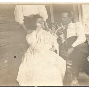 May include: A black and white vintage photograph of three people sitting on a porch. A woman in a white dress is sitting in the middle, with a man in a white shirt and dark pants sitting to her right. A woman in a white blouse and dark skirt is standing behind the woman in the white dress.