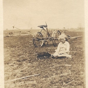May include: A black and white photo of a young girl sitting in a field with a farm implement in the background. The girl is wearing a light colored dress and has her hair pulled back. The implement has a large wheel and a seat.