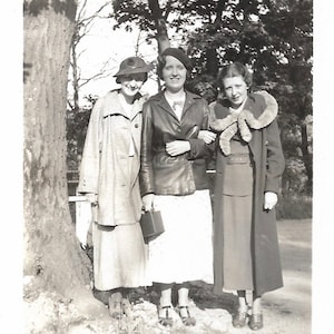 May include: Three women standing in front of a tree. The woman on the left is wearing a light-colored coat and a hat. The woman in the middle is wearing a dark leather jacket and a hat. The woman on the right is wearing a dark coat with a fur collar.