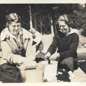 May include: Two women wearing sweaters and skirts are sitting on a wooden dock by a lake. One woman is holding a bucket and the other is holding a jar. The women are smiling and appear to be enjoying their time together.