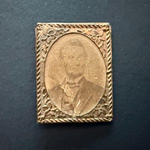 May include: A small, rectangular antique photograph in a gold-colored metal frame. The frame has an oval opening and is decorated with floral and rope-like patterns. The photograph features a sepia-toned portrait of a person.