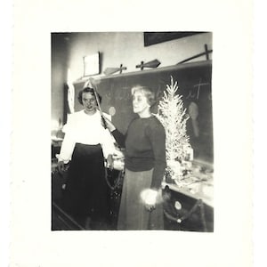 May include: Black and white photo of two women standing in front of a decorated Christmas tree. The woman on the left is wearing a white blouse and a dark skirt. The woman on the right is wearing a dark sweater and a skirt. The tree is decorated with white ornaments and a star on top.