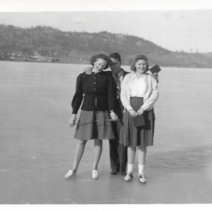 May include: Three people, two women and a man, are standing on a frozen lake. The women are wearing skirts and jackets, and the man is wearing a jacket and pants. The women are holding purses.