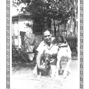 May include: A black and white photo of a woman with two dogs. The woman is smiling and holding one dog in her arms. The other dog is standing next to her. The photo is framed with a decorative border.