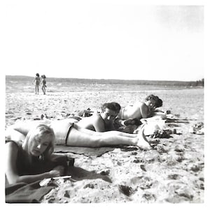 May include: Black and white photograph of several people sunbathing on a sandy beach. Two children are in the distance near the water. The image captures a moment of leisure and relaxation.
