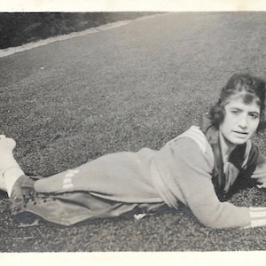 May include: Vintage black and white photograph of a person lying on the grass. The person is wearing a sweater, pants, and shoes. The photograph is taken outdoors on a sunny day.