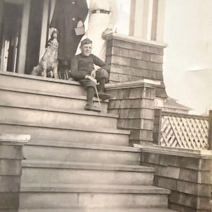 May include: Vintage black and white photograph of three people and a dog on a porch. A young person sits on the steps, holding a cane. Two adults stand behind them. The dog is sitting on the porch.