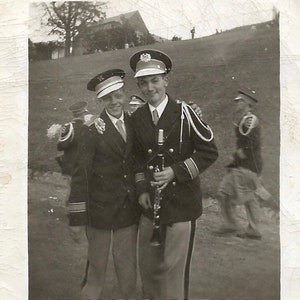 May include: Black and white photograph of two young men in band uniforms, posing outdoors. One holds a clarinet. They wear hats with insignia, dark jackets, light-colored pants, and ties. Other band members are visible in the background.