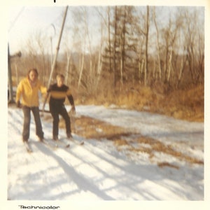 May include: Two people in winter clothing are skiing down a snowy slope. The person in the front is wearing a yellow jacket and the person in the back is wearing a black jacket. The slope is covered in snow and there are trees in the background.