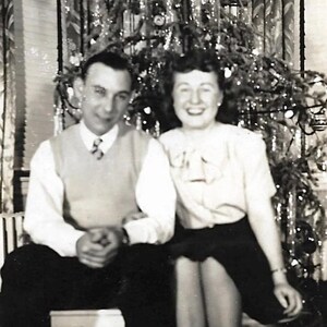 May include: A black and white photo of a couple standing in front of a Christmas tree. The man is wearing a white shirt and a sweater vest. The woman is wearing a white blouse and a dark skirt. They are both smiling.