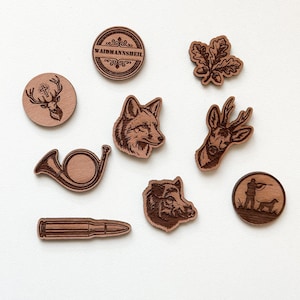 Könnte beinhalten: Eine Sammlung von neun laser-gravierten Holz-Anstecknadeln mit verschiedenen Jagdmotiven, darunter ein Hirsch mit Kreuz, ein Fuchs, ein Wildschwein, ein Jagdhorn, eine Patrone, ein Eichenblatt, ein Jäger mit Hund sowie ein rundes Waidmannsheil-Emblem.