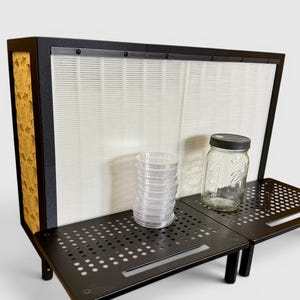 May include: A black laminar flow hood featuring a large white pleated filter, a side panel with a gold-toned pattern, and two black perforated metal work surfaces. A stack of clear plastic petri dishes and a glass jar with a black lid sit on top.