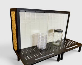 May include: A black laminar flow hood featuring a large white pleated filter, a side panel with a gold-toned pattern, and two black perforated metal work surfaces. A stack of clear plastic petri dishes and a glass jar with a black lid sit on top.