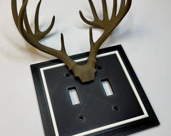 May include: A decorative light switch cover featuring a black plate with a white border and two switch openings. The cover is topped with a brown antler design, adding a rustic touch. The design is suitable for a home or cabin.