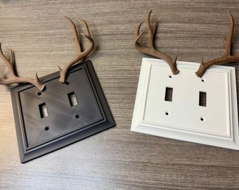 May include: Two decorative light switch covers, one black and one white, each adorned with faux brown deer antlers. The covers have two switch openings and are set against a wood-grain background.