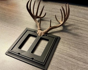 May include: A decorative light switch cover featuring a brown antler design. The cover is black and has two rectangular openings. The antlers are a light brown color and protrude from the top of the cover.