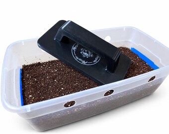 May include: A clear rectangular plastic container filled with dark brown soil, featuring blue tape along the inner edges and circular side holes. A black rectangular lid with a central handle and a circular white logo reading MycoAir sits on top.
