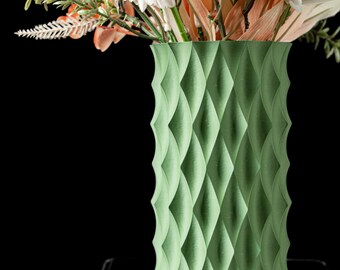May include: A green vase with a geometric diamond pattern, filled with a bouquet of artificial flowers. The flowers include peach-colored dahlias and white blossoms, with green and brown foliage. The vase is cylindrical and sits on a reflective surface.