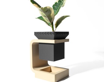 May include: A modern plant stand with a square black planter holding a green and white variegated plant. The stand has a light beige C-shaped support and a dark gray cube beneath the planter.
