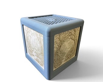 May include: A matte blue cube-shaped container with a textured surface and a honeycomb-patterned lid. The side panels feature detailed, light-colored illustrations of a forest landscape with trees and foliage, set within a recessed frame.
