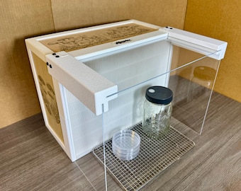 May include: A white laboratory still air box with clear acrylic panels, featuring a top panel with mushroom illustrations and the text Mycoho. Inside, a metal wire rack holds a glass jar with a black lid and a stack of clear plastic petri dishes.