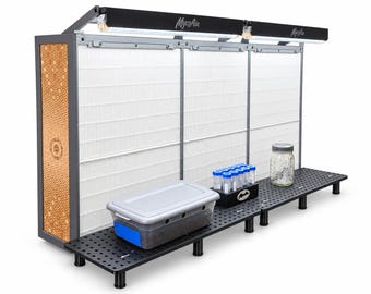 May include: A MycoAir laminar flow hood featuring a large white HEPA filter, a black frame with a decorative honeycomb side panel, and an integrated LED light bar. A black perforated work surface holds a plastic storage bin and glass jars.
