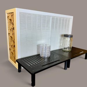 May include: A white air filtration unit with a side panel featuring a brown mushroom pattern, positioned behind two black perforated metal monitor stands. A stack of clear plastic petri dishes and a glass jar with a lid sit on the stands.