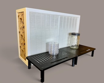 May include: A white air filtration unit with a side panel featuring a brown mushroom pattern, positioned behind two black perforated metal monitor stands. A stack of clear plastic petri dishes and a glass jar with a lid sit on the stands.
