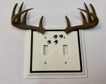 May include: A decorative light switch cover featuring a pair of brown deer antlers. The cover is white with a black border and includes two toggle switch openings and a set of black deer tracks.