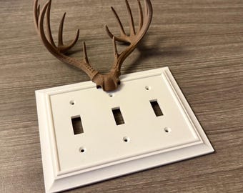 May include: A white triple light switch cover with a decorative brown antler design. The cover has three switch openings and four screw holes. The antler design is mounted above the switch plate.