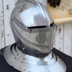 May include: A polished steel knight's helmet with intricate floral and geometric etched designs. The helmet features a visor, a neck guard with rivets, and a decorative crest. The metal has a reflective surface.