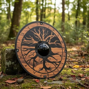 Pode incluir: Um escudo redondo de madeira com um desenho de árvore da vida preta no centro. O escudo tem um padrão de grão de madeira marrom escuro e uma borda de metal preta. Está sobre uma pedra coberta de musgo em um ambiente florestal.