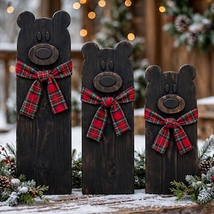 May include: Three dark brown wooden bear-shaped cutouts of varying heights, each featuring a rounded snout, two small oval eyes, and a red, green, and yellow plaid fabric ribbon tied around the neck as a decorative scarf.