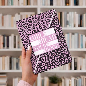 Op de afbeelding: Een roze en zwarte luipaardprint boekomslag met de tekst "GIRL READ YOUR BOOK" in een witte rechthoekige doos. De omslag wordt in een hand gehouden, met een boekenplank op de achtergrond.