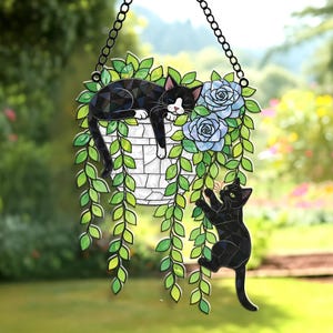 May include: A decorative hanging stained glass cat planter with a sleeping black and white cat, blue roses, and a climbing black cat. The planter is white and the leaves are green. The piece hangs from a black chain.