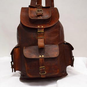 May include: A brown leather backpack with multiple pockets and brass buckles. The backpack has a flap closure on the top and two side pockets. The leather has a distressed appearance.