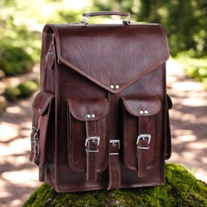 May include: A brown leather backpack with multiple pockets and silver buckles. The bag has a top handle and a flap closure. The backpack is sitting on a mossy surface, with a blurred green background.