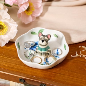 May include: A white ceramic trinket dish with a scalloped edge, featuring painted green leaves and blue floral accents. A small, hand-painted ceramic dog figurine wearing a green sweater sits in the center, surrounded by four silver-toned rings.