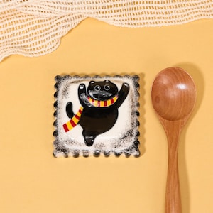 May include: A square ceramic dish with a black cat design. The cat is black with a yellow and red scarf, arms raised in a joyful pose. The dish has a white background and a scalloped black border. A wooden spoon rests beside it.