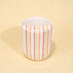 Könnte beinhalten: Eine weiße Keramiktasse mit vertikalen Streifen in Rot, Gelb und Türkis. Die Streifen sind gleichmäßig verteilt und verlaufen vom Boden bis zum Rand der Tasse. Das Innere der Tasse ist weiß.