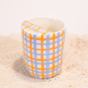 May include: A ceramic toothbrush holder with a blue and orange plaid pattern. The holder has a curved handle and a white interior. The design is a grid of intersecting lines in blue and orange.