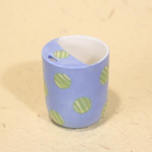 May include: A cylindrical ceramic toothbrush holder. The holder is light blue with green and white striped polka dots. The interior is white. The top has a curved design with a small hole.