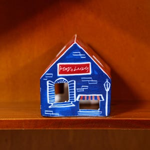 Könnte beinhalten: Eine kleine blaue Keramikhausfigur mit rotem Dach. Die Vorderseite zeigt handgemalte weiße Details, darunter ein Fenster mit Fensterläden, eine gestreifte Markise über einem Eingang, eine Straßenlaterne und ein rotes Schild mit weißer Schrift.