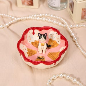 May include: A ceramic trinket dish shaped like a bowl with a red drip rim, pink interior, and waffle cone patterns. A small black and white cat figurine with a pink collar and bell sits in the center. The name Suki is written in black on the base.