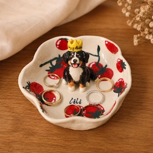 May include: A cream-colored ceramic trinket dish with a scalloped edge, decorated with red tomato illustrations. A small, three-dimensional Bernese Mountain Dog figurine wearing a yellow crown labeled Happy Birthday sits in the center, with the name lili.