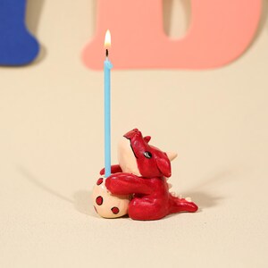 Pode incluir: Uma figura de dragão vermelho segurando uma vela azul clara com uma chama acesa. O dragão tem detalhes brancos e abraça uma bola bege com manchas vermelhas. O fundo inclui uma letra azul e rosa.