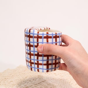 May include: A ceramic mug with a blue and brown plaid pattern. The mug is cylindrical with a small handle and is held by a hand. The interior of the mug is white.
