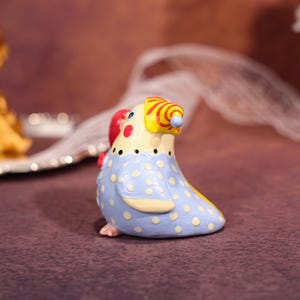 Op de afbeelding: Een keramieken vogelbeeldje met een grillig ontwerp. De vogel heeft een lichtblauw lichaam met witte stippen, een gele kop met rood-gele strepen en een rood hartvormig accent. Het beeldje is ongeveer 7.6 cm hoog.